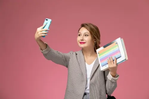 Jovem aluna com casaco cinza posando segurando livros com um sorriso tirando uma selfie